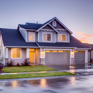 Modern gray two-story house with illuminated windows and a wet driveway reflecting the sunset colors.