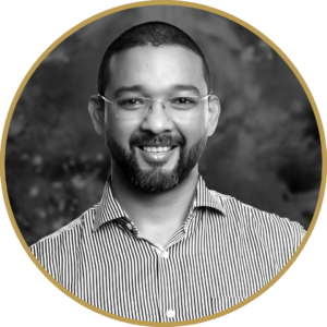 Black and white professional headshot of Delano, a smiling man with a beard and glasses wearing a striped shirt.